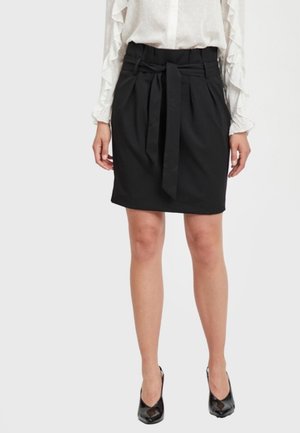 Femme portant une jupe noire arrivant aux genoux avec une ceinture nouée, associée à des talons noirs et un chemisier blanc à manches longues avec des manches à volants.