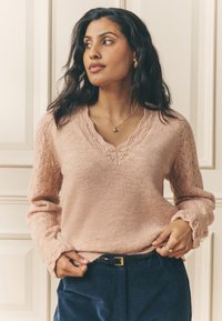 Różowy sweter z dzianiny z dekoltem w serek i koronkowymi zdobieniami na rękawach; w połączeniu z granatowymi sztruksowymi spodniami i czarnym paskiem. Prosty złoty naszyjnik.