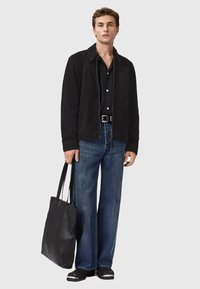 Mand iført sort ruskindsjakke, sort knappet skjorte, brede blå jeans, sorte sko med hvide spidser, bærende en stor sort læder tote-bag.