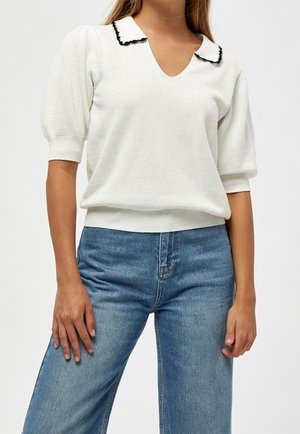 Femme portant un pull à manches courtes blanc texturé avec un col bordé de noir et un jean bleu taille haute, sur un fond uni.