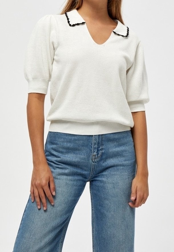 Femme portant un pull à manches courtes blanc texturé avec un col bordé de noir et un jean bleu taille haute, sur un fond uni.