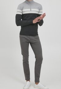 Mann trägt einen schwarz, grau und weiß farblich abgesetzten Pullover, graue Slim-Fit-Hosen und weiße Sneakers und steht vor einem einfarbigen Hintergrund.