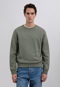 Olive green sweatshirt with ribbed neckline and cuffs, featuring a subtle logo on the chest. Worn over a white shirt, paired with blue jeans.