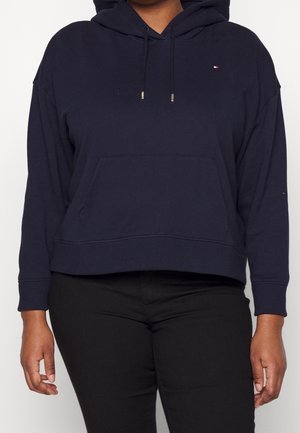 Person wearing a navy blue hoodie with front pocket and small logo on chest, paired with black pants, against a plain background.