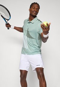 Polo sportivo verde con motivi astratti, abbinato a pantaloni corti bianchi. Il modello tiene in mano una pallina da tennis e una racchetta, mostrando abbigliamento sportivo.