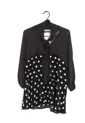 Blouse noire avec des pois blancs en haut et des motifs de cœurs blancs en bas, avec un nœud noué au cou, accrochée à un cintre noir.