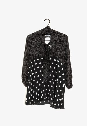 Blouse noire avec des pois blancs en haut et des motifs de cœurs blancs en bas, avec un nœud noué au cou, accrochée à un cintre noir.