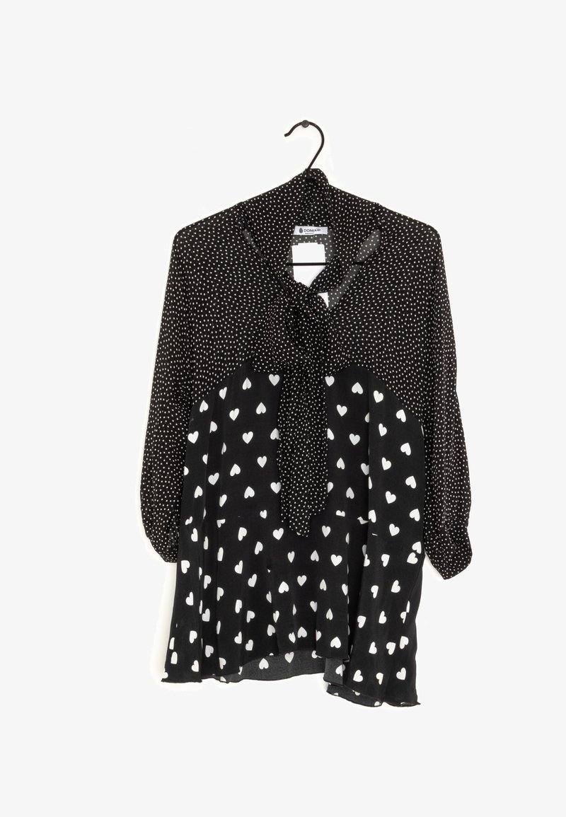 Blouse noire avec des pois blancs en haut et des motifs de cœurs blancs en bas, avec un nœud noué au cou, accrochée à un cintre noir.