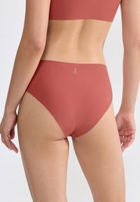 Donna che indossa lingerie senza cuciture di colore corallo scuro con vita alta e reggiseno abbinato, mostrata di spalle su uno sfondo semplice e chiaro.