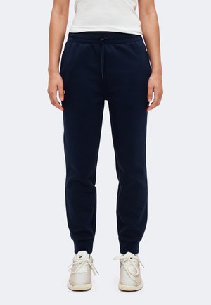 Un modèle porte un pantalon de jogging bleu marine à cordon de serrage, resserré aux chevilles, ainsi que des baskets blanches sur un fond clair uni.