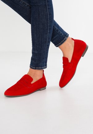 Personne portant un jean en denim bleu et des mocassins en daim rouge vif, debout sur une surface blanche et lisse.