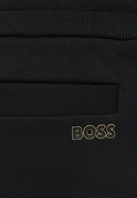 Čierna látka s rebrovanou textúrou, horizontálnym vreckom a zlatým logom "BOSS". Jednoduchý dizajn s hladkým povrchom.