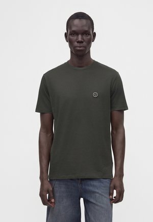Homme portant un t-shirt à manches courtes vert foncé avec un petit logo rond sur la poitrine, associé à un jean bleu, debout devant un fond uni.