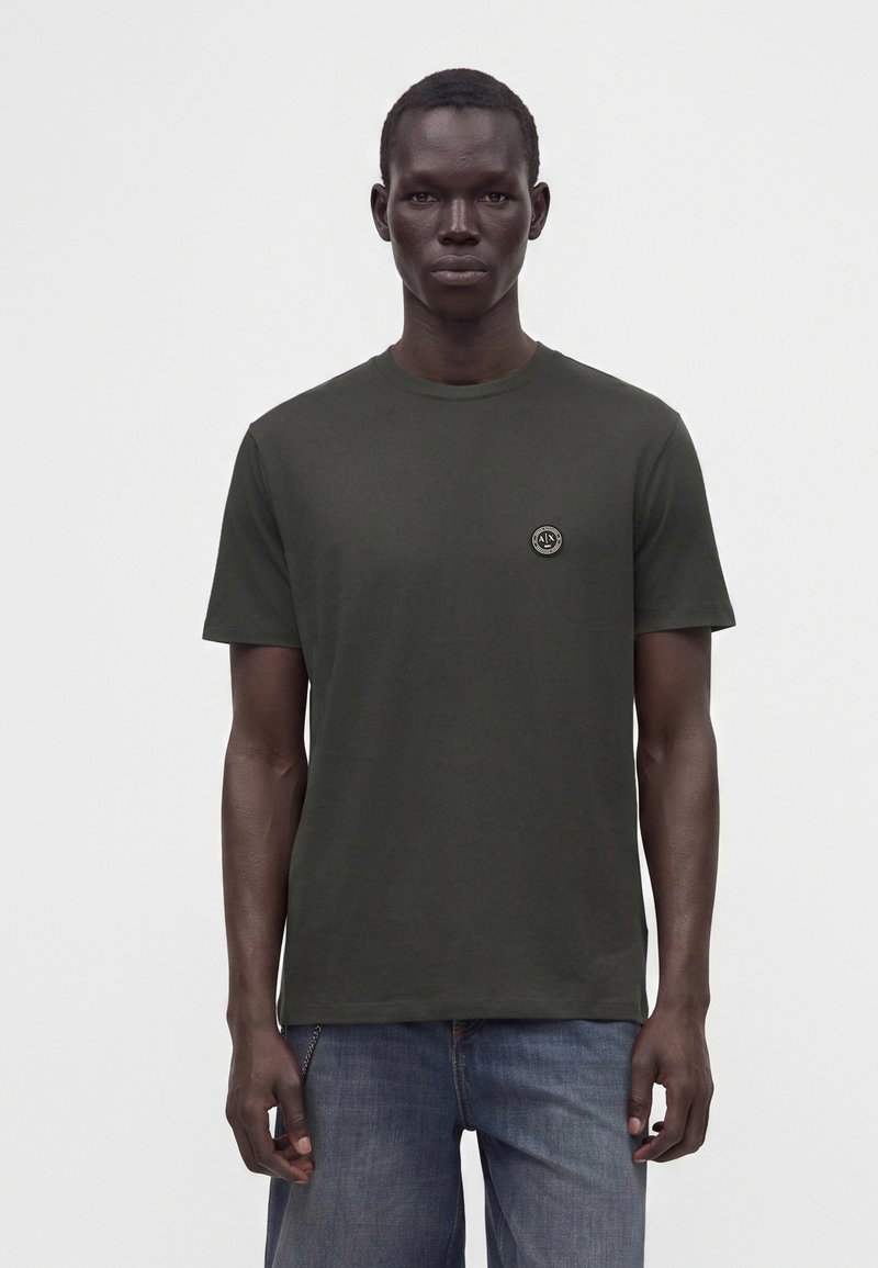 Homme portant un t-shirt à manches courtes vert foncé avec un petit logo rond sur la poitrine, associé à un jean bleu, debout devant un fond uni.