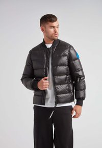 Giacca a vento nera con chiusura a zip, polsini a costine e un accento blu sulla manica sinistra. Presenta un design trapuntato e una finitura lucida.