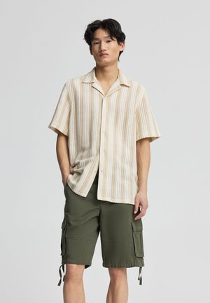 Giovane uomo in piedi con le mani nelle tasche, indossa una camicia beige a righe a maniche corte e pantaloncini cargo verde oliva su sfondo uniforme.