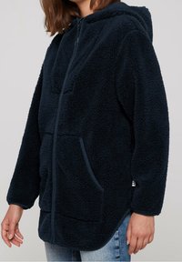 Mörkblå fleecehuva med dragkedja, stora framfickor och en texturerad yta. Har en huva och långa ärmar med muddar.