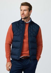 Man draagt een marineblauwe doorgestikte bodywarmer over een gebrande oranje trui en marineblauwe broek, kijkt iets naar rechts met ontspannen handen.