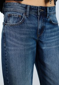 Tamnoplave traperice ravnog kroja, sa vidljivim štepom, dva prednja džepa i klasičnim zatvaračem na gumbu u struku.