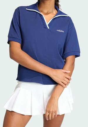 Persona con una camiseta polo azul de manga corta con ribete blanco y una falda plisada blanca, con los brazos cruzados sobre el cuerpo.