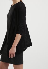Cardigan negro, ligero, con mangas largas, de frente abierto y diseño fluido, superpuesto a un vestido oscuro con textura.