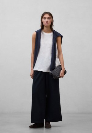 Mladá žena stojící opřená o šedou stěnu, má na sobě bílý top bez rukávů, tmavě modré široké kalhoty, přes ramena přehozený tmavě modrý svetr a drží tmavé psaníčko.