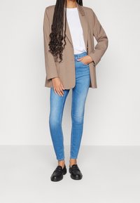 Mujer con blazer marrón claro, camiseta blanca, jeans ajustados azul claros, mocasines negros y cabello largo trenzado, contra un fondo simple.