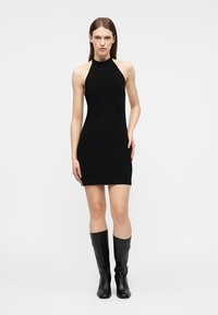 Γυναίκα που στέκεται, φορώντας μια μαύρη μίνι φόρεμα χωρίς μανίκια με halter neck και μαύρες δερμάτινες μπότες μέχρι το γόνατο, πάνω σε απλό λευκό φόντο.