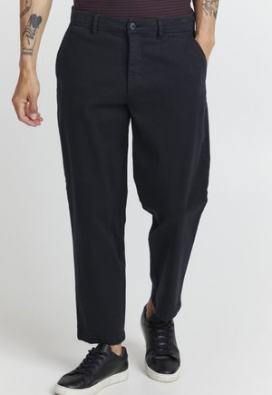 Black cotton trousers with a straight fit, featuring side pockets and a button closure. Worn with black sneakers, displaying a relaxed style.