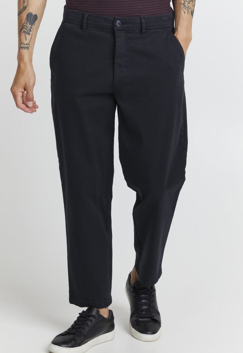Pantalon en coton noir avec une coupe droite, doté de poches latérales et d'une fermeture à bouton. Porté avec des baskets noires, affichant un style décontracté.