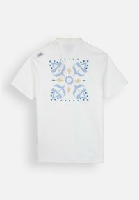 Polo shirt blanc à manches courtes avec un design graphique floral bleu et beige au dos, accompagné d'une étiquette de logo sur le col. Fabriqué en coton.