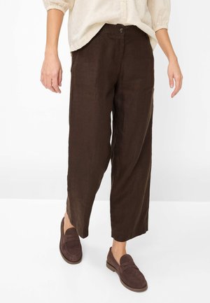 Vrouw draagt donkerbruine, brede, cropped broek, bijpassende bruine loafers en een crèmekleurige blouse met driekwartmouwen.