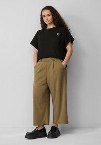 T-shirt noir à manches courtes avec un petit logo blanc, associé à un pantalon beige à jambes larges avec deux boutons. Chaussures à plateforme noires.