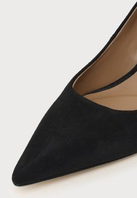Chaussure pour femme en daim noir à bout pointu avec une doublure intérieure en cuir beige, présentée sur un fond clair.