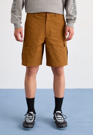 Man draagt bruine cargoshorts, zwarte sokken en zwart-witte sneakers op een blauwe vloer tegen een witte muur.
