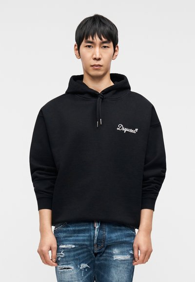 Μαύρο φούτερ με μεγάλο τσέπη καγκουρό, κορδόνια στη κουκούλα και λογότυπο «Dsquared2» με λευκή κεντητική. Φορεμένο με ξεφτισμένο τζιν παντελόνι.