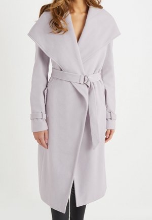 Femme portant un long manteau gris clair ceinturé avec un large col et des détails de boucles sur les manches sur un fond blanc uni.