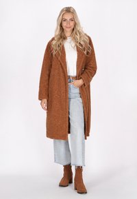 Brun texturerad oversizekappa med framknapp, bärs över en vit skjorta, tillsammans med ljusblå vida jeans och bruna klackstövlar.