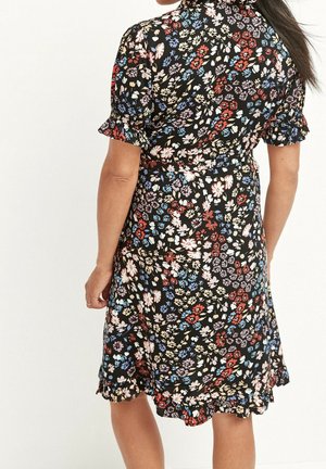 Robe noire à imprimé floral rouge, bleu et crème, manches courtes bouffantes et ourlet à volants, portée par une personne vue de dos.