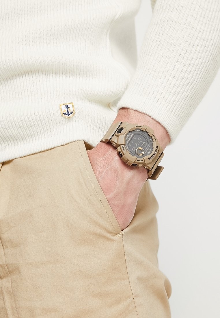 Personne portant un pull en maille blanc et un pantalon beige, une main dans la poche, montrant une montre digitale G-Shock beige au poignet.