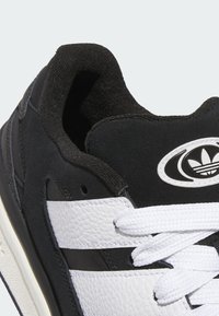 Zapatilla negra con parte superior de gamuza negra texturizada, franjas de cuero blanco, cuello acolchado y cordones blancos con un acento de logo.