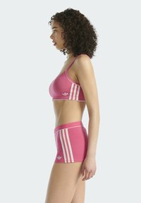 Růžový sportovní komplet obsahující podprsenku a odpovídající šortky. Materiál je hladký s bílými pruhy a logem po stranách.