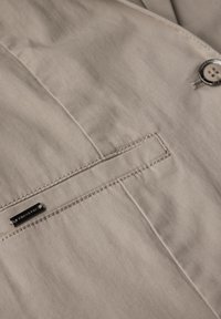 Beige tyg med en slät textur, med en enda bröstficka med sydda kanter och en rund vit knapp med en svart kant.