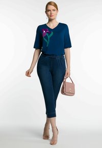 Navy blue V-necka paita, jossa on violetteja ja vihreitä kukka-ompeluksia, yhdistetty tummansinihin nilkkahousuihin ja vaaleanpunaiseen käsilaukkuun, jossa on kultainen solki.