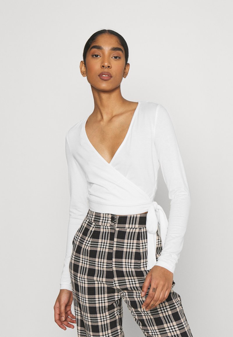 Blusa branca de manga longa em estilo envolvente com um decote em V profundo, combinada com calças de xadrez preto e branco de cintura alta com um detalhe de laço na cintura.