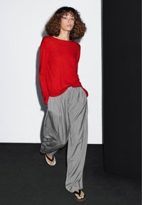 Haut rouge à manches longues, pantalon ample gris plissé et tongs noires. Textures de tissu lisses et silhouette décontractée avec un drapé subtil.