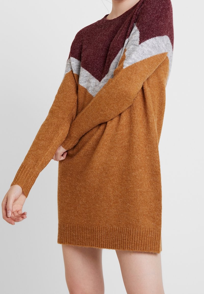 Femme portant une robe-pull longue bicolore marron, grise et bordeaux, ajustant une manche avec sa main opposée.