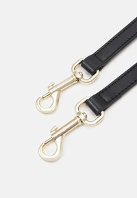 Deux lanières en cuir noir chacune équipée d’un clip pivotant en métal doré, les clips ont des crochets à bouton-poussoir sur un fond clair uni.