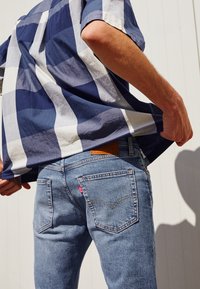 Blå och vit rutig skjorta över blå denimjeans med en synlig Levi's-logotyp och sömnadsdetaljer på bakfickorna.