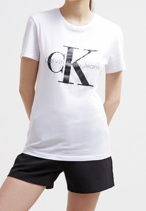 Femme portant un t-shirt blanc Calvin Klein Jeans avec un grand logo noir "ck" et un short noir, debout les mains derrière le dos.